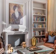 Elegant living room with two armchairs, a fireplace, and built-in bookshelves. A painting hangs above the fireplace, and a chess set is on a glass coffee table in the center.