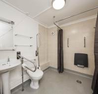Accessible bathroom with a toilet, sink, and multiple grab bars. Dark curtains around shower area. Mirror and shelves above the sink. Tiled walls and non-slip flooring.