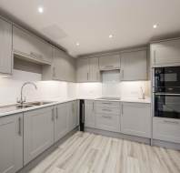Modern kitchen with gray cabinets, stainless steel sink, and built-in appliances including an oven and microwave. Wood-like flooring and under-cabinet lighting highlight the space.