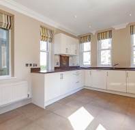 Modern kitchen with beige walls, tiled floor, and white cabinets. Features large windows with striped blinds and a dark countertop with a sink. Radiator beneath one window.