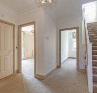 Hallway with beige carpet, two closed doors, a room with curtains, and a carpeted staircase leading up. A large window at the stair top lets in natural light.