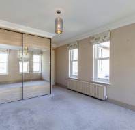 Empty room with beige walls, carpeted floor, mirrored wardrobe, two windows with patterned blinds, and a ceiling light fixture.