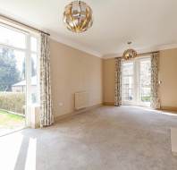 Spacious, carpeted living room with large windows, patterned curtains, two pendant lights, a wall-mounted TV, and a decorative fireplace. Glass doors lead to an outdoor view.