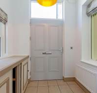 Entrance hallway with a closed gray door, sidelights, and a window above. Beige tiled floor, radiator on the right, and wooden cabinets on the left. Ceiling features a yellow hanging light.