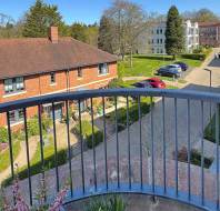 View from a balcony overlooking a courtyard with brick buildings, greenery, and parked cars on a sunny day.