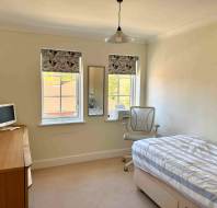 Minimalist bedroom with a single bed, striped bedding, desk with a monitor, chair, and two windows with floral blinds. A mirror is between the windows.