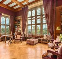 A spacious, elegant library with tall windows, purple drapes, wooden floors, bookshelves, and comfortable seating.