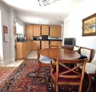 A dining area with a wooden table and chairs, a glass cabinet, a patterned rug, and an adjoining kitchen with light wood cabinets and appliances.