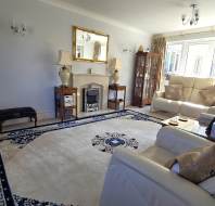 A bright living room with cream sofas, a patterned rug, TV, display cabinet, fireplace, large window, and various lamps and ornaments.