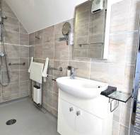 Accessible bathroom with a walk-in shower, wall-mounted shower seat, grab bars, sink with cabinet, mirror, and a heated towel rail on tiled walls.