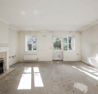 Empty, unfurnished room with a fireplace, two windows, two radiators, and worn carpet. Sunlight enters through the windows, casting shadows on the floor.