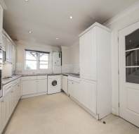 A modern kitchen with white cabinets, built-in ovens, a washing machine, and a window above the sink, viewed from the entrance door.