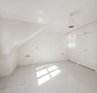 Empty white room with built-in wardrobes, a window letting in natural light, radiator under the window, and sloped ceiling on one side.