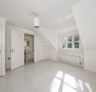 Unfurnished white attic room with sloped ceiling, radiator, window, open door leading to a small sink area, and another closed door. Sunlight streams in through the window.