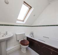 White tiled bathroom with pedestal sink, toilet with wooden seat, bathtub with shower, and a skylight window in a sloped ceiling.