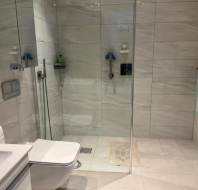 Modern bathroom with large tiled shower, glass partition, wall-mounted toilet, sink, towel rack with a folded towel, and a floor mat inside the shower area.