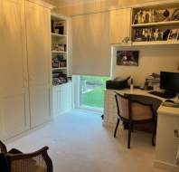 Home office with built-in shelves, a desk with a computer and printer, a window with a shade, two chairs, and various photos and memorabilia on display.