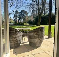 View through a glass door showing two wicker chairs and a round table on a patio, overlooking a grassy garden with trees under a clear blue sky.