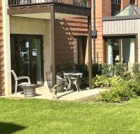 A small patio with wicker chairs, a table, and a bird sculpture outside a brick building, next to a windowed extension, bordered by green lawn and shrubs.