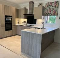 Modern kitchen with light wood cabinets, built-in appliances, white countertops, a sink in the island, and a floral patterned window blind.