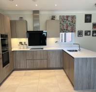 Modern kitchen with light wood cabinets, a built-in oven, electric cooktop, stainless steel range hood, white countertops, and a small sink; floral blind on window and photos on the wall.