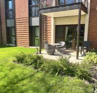 A ground-floor patio with two wicker chairs and a table, partially shaded by a balcony, overlooks a grassy lawn next to a brick apartment building.