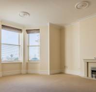 Empty room with beige walls, three large bay windows with blinds, beige carpet, white trim, and a small electric fireplace on the right wall.