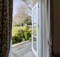 An open glass door with patterned curtains reveals a stone patio and a green garden with trees and hedges in daylight.