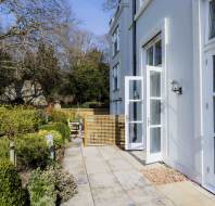 A white exterior door stands open onto a paved patio with outdoor furniture, surrounded by a neatly landscaped garden and greenery.