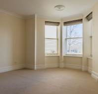 Empty beige room with a bay window, white trim, and tan carpet, lit by natural light coming through multiple windows with blinds.