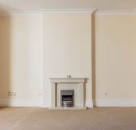 A spacious, empty room with beige walls, cream carpet, and a central fireplace beneath a mantel, with a white ceiling and trim.
