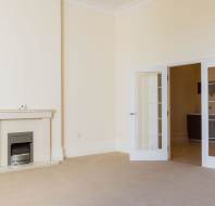 Spacious empty room with beige carpet, cream walls, a fireplace on the left, and white double glass doors leading to another area.