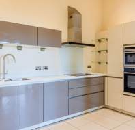 Modern kitchen with glossy brown and beige cabinets, built-in oven and microwave, stainless steel sink, under-cabinet lighting, and tiled floor.