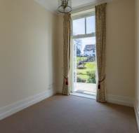 Small, empty room with beige carpet and cream walls, featuring a tall window with patterned curtains overlooking a garden and a house outside.