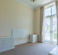 Empty bedroom with cream walls, two white dressers, a headboard, large window with vertical blinds, curtains, and a chandelier.