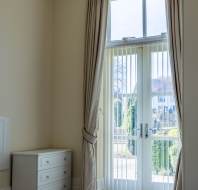 A tall window with vertical blinds and beige curtains beside a white chest of drawers in a bright, empty room.