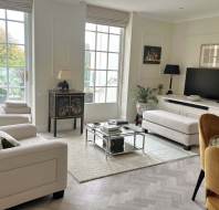 Bright, modern living room with large windows, neutral furniture, a TV on a console, glass coffee table, and art on the walls.