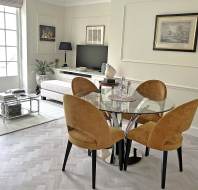 A modern living and dining area features a glass dining table with four tan chairs, a white sofa, TV, and large window, all set on light herringbone flooring.