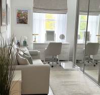 A tidy, light-filled room with a white sofa, glass coffee table, and a desk with two chairs facing a window; mirrored closet doors reflect the space.