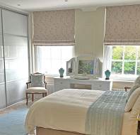Bright bedroom with a large bed, vanity table, chair, and big windows with patterned blinds. Sliding closet doors are on the left. The decor is light and neutral.