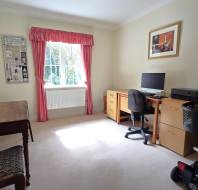 A home office with a desk, computer, printer, two chairs, a mobility scooter, and a window with pink curtains letting in natural light.