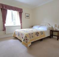 A neatly made bed with a patchwork quilt is placed beside a window with pink curtains in a simply furnished bedroom with cream walls and carpet.