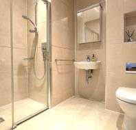Modern bathroom with beige tiles, a glass-enclosed shower, wall-mounted sink, mirror, toilet, and toiletries neatly arranged.