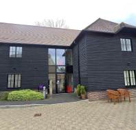 A two-story building with dark wooden siding, a glass entryway, brick trim, benches, and landscaped greenery in front.