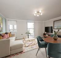 Modern living room with a white sectional sofa, colorful cushions, a round wooden dining table with teal chairs, a large wall mirror, and a TV on a console. Large window lets in natural light.