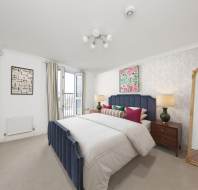 A well-lit bedroom with a double bed, colorful pillows, a large mirror, wall art, a wooden dresser with a vase, and a ceiling light fixture, featuring a partial view of a window and balcony.