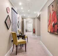 A hallway with beige walls and carpet, featuring a small round table and two yellow chairs. Wall art, including a portrait and geometric decorations, hang along the left wall.