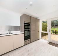 Modern kitchen with light wood cabinets, built-in ovens, and a sink. Glass doors open to a patio with outdoor dining set and grassy garden area. Bright and minimalist design.