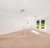 An empty, bright room with beige carpet, white walls, ceiling lights, double doors, and a small window. Natural light enters through the doors, revealing a view of a green garden.