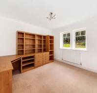 Room with a beige carpet, wooden desk, and shelves. A white wall has two large windows, and a small ceiling light fixture is visible.
